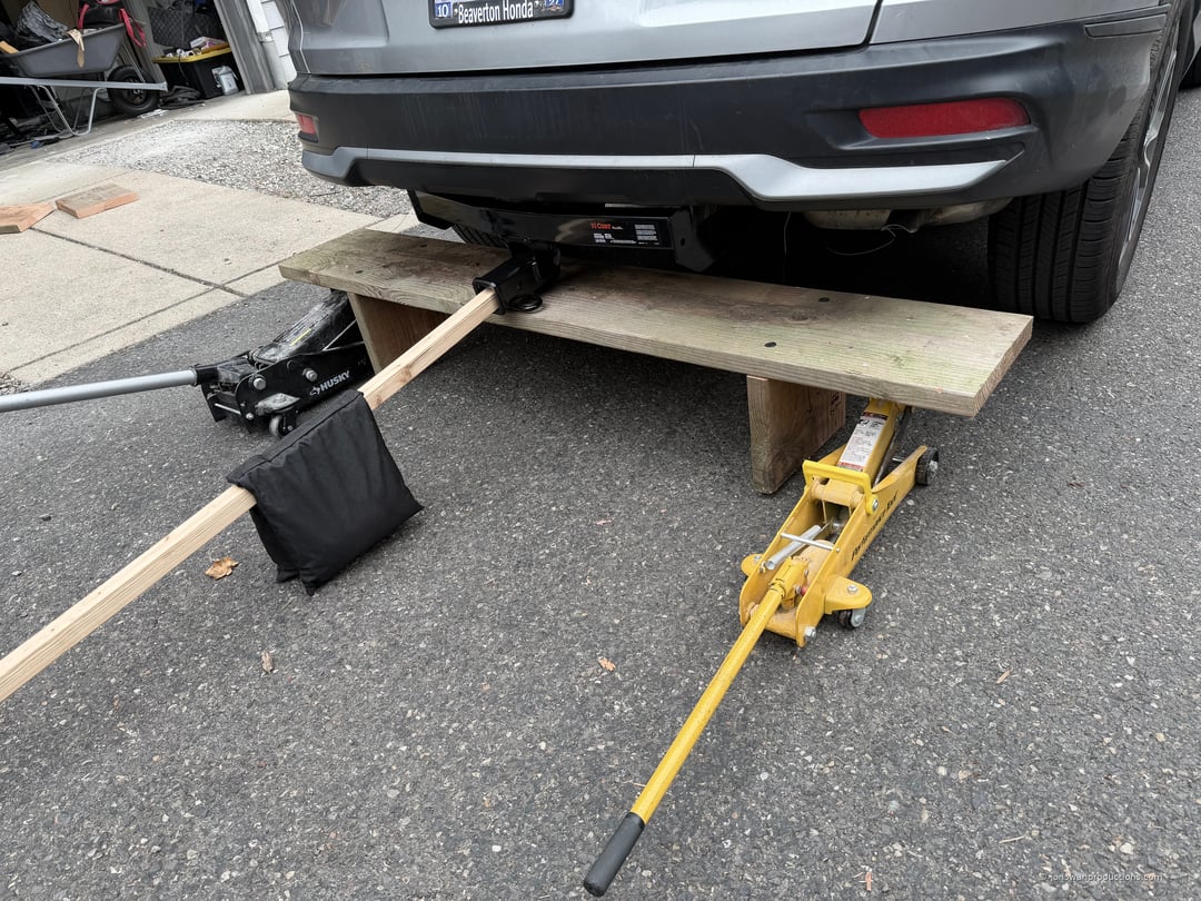 Two floor jacks supporting a shared plank under a Honda CR-V, holding the CURT hitch in position, with a lever arm and sandbag counterweight